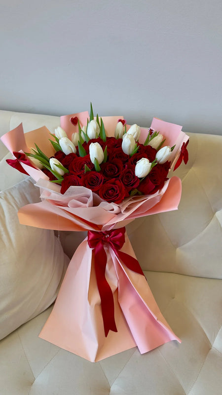 Amour Signature 50 bouquet with red roses and white tulips, wrapped in blush pink paper and finished with a red satin bow.