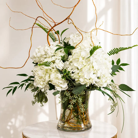 White hydrangeas with greenery and curly willow branches arranged in a clear glass vase.