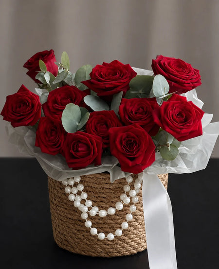 Red roses and eucalyptus in a woven basket decorated with white pearl accents and a white ribbon.