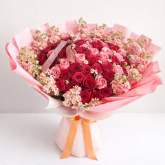 Simple Dulzura bouquet with red and pink roses, pink stock and small white chamomile flowers, wrapped in pink paper with a peach ribbon.