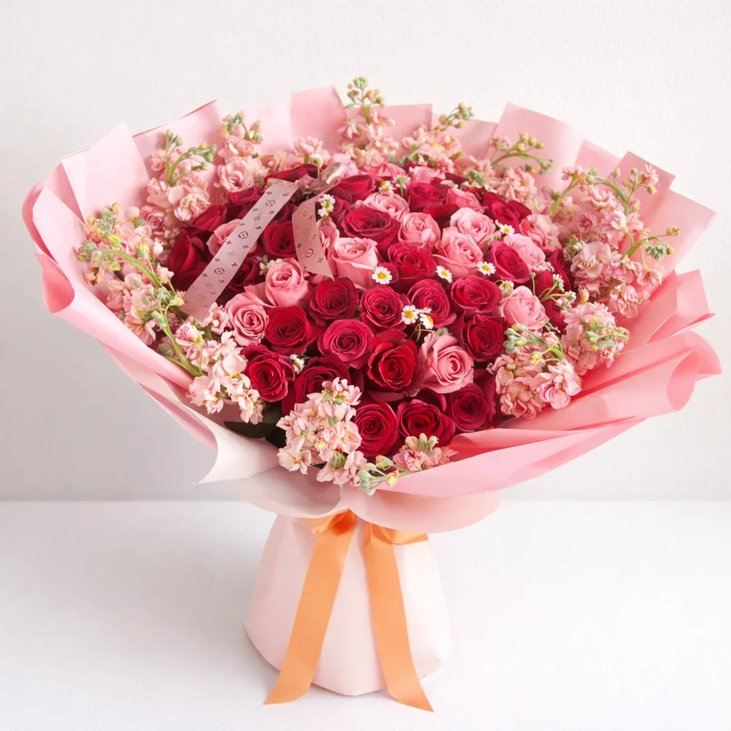Simple Dulzura bouquet with red and pink roses, pink stock and small white chamomile flowers, wrapped in pink paper with a peach ribbon.
