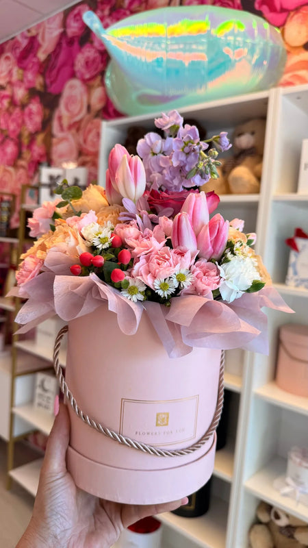 Pink tulips, roses, carnations and seasonal flowers arranged in a round pink flower box.